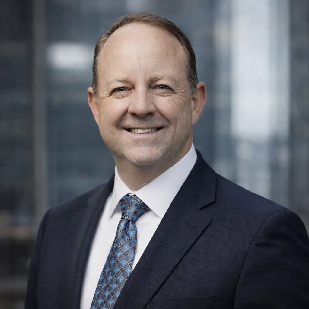 Smiling man in a navy suit and blue patterned tie against a blurred office background.