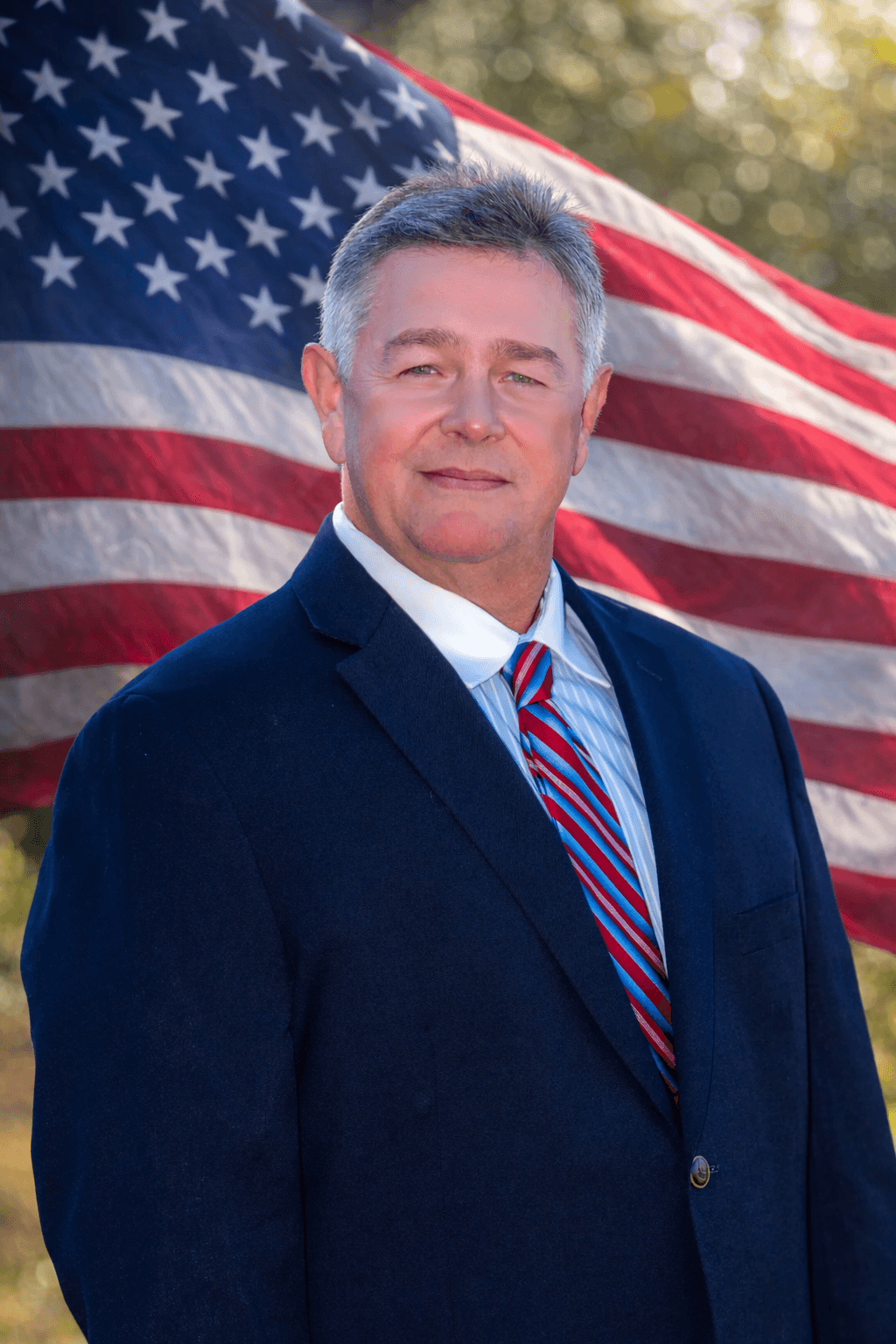 Middle-aged man in a navy suit and striped tie posing before an American flag.