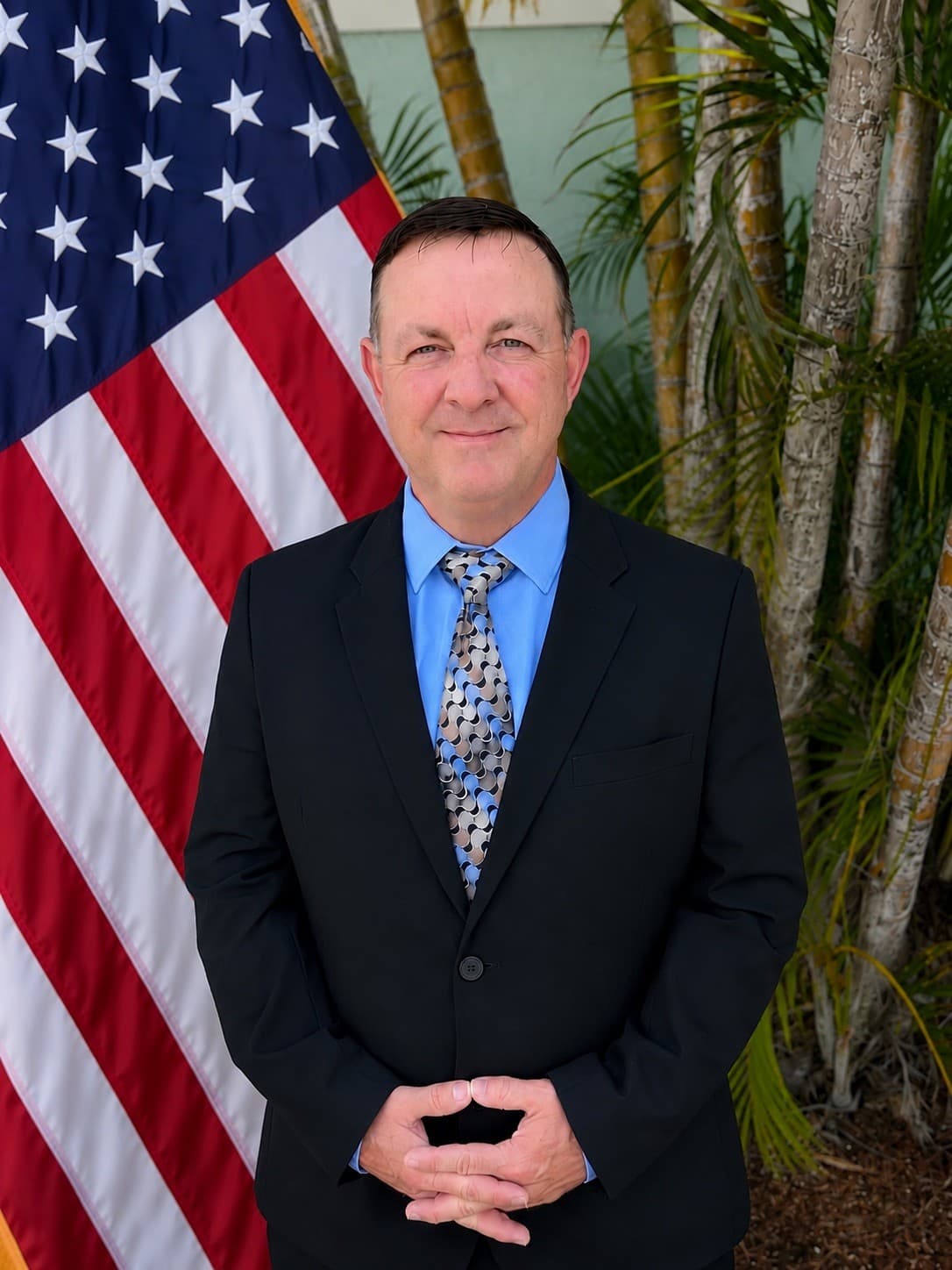 Man in a suit and tie stands before an American flag and palm trees.